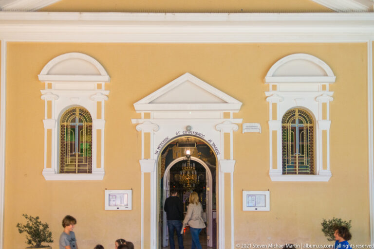 ochre church façade with white trim, two arched windows, central doorway, and visitors entering.