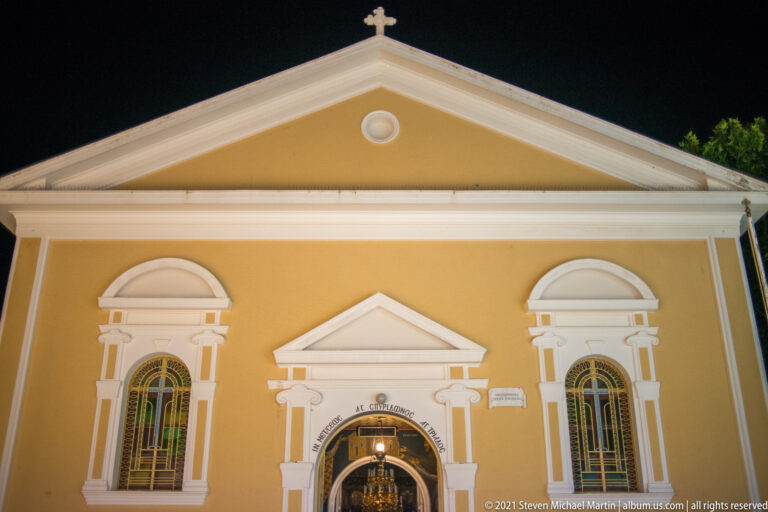 Upper ochre church façade at night with white pediment, cross, paired arched windows, and central entrance below.