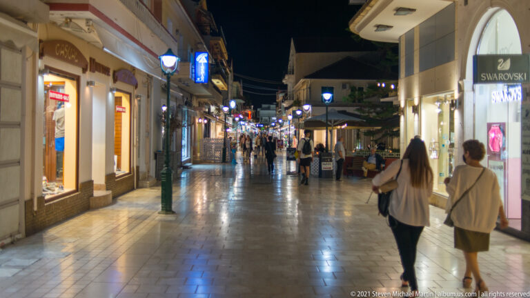 Pedestrians walking along a brightly lit nighttime shopping street with boutiques, cafés, and outdoor seating.