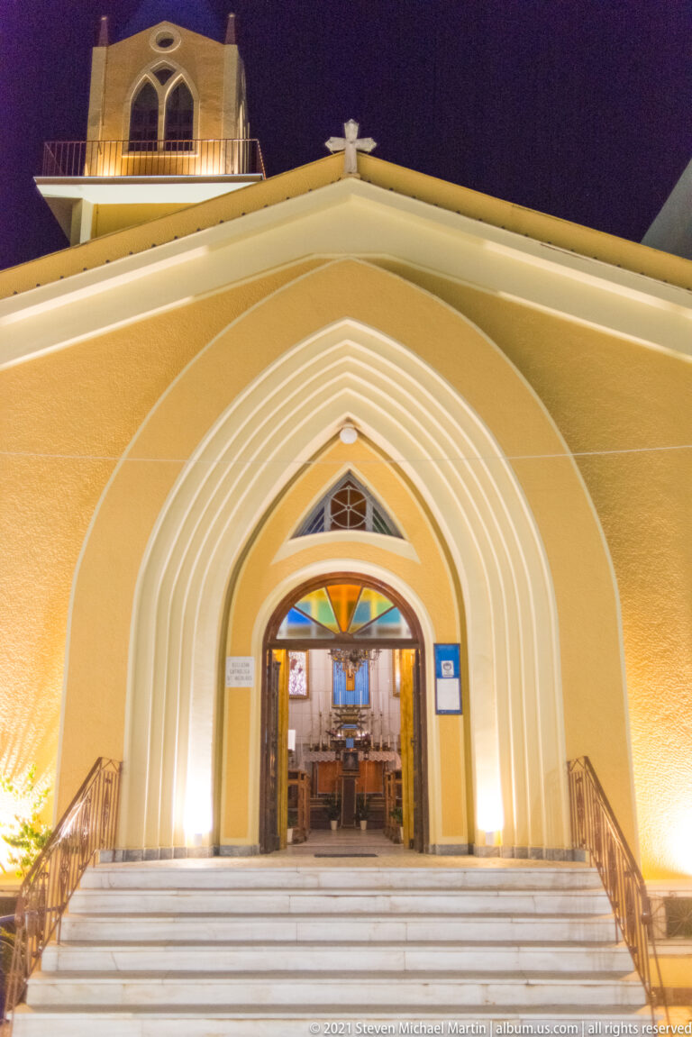 Illuminated church entrance with broad white steps, nested pointed arches, open doors, and colored glass above.