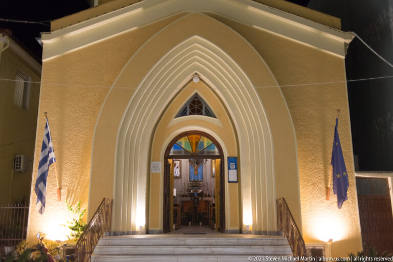 Floodlit pointed-arch church entrance with Greek and EU flags flanking steps and open doors to the interior.