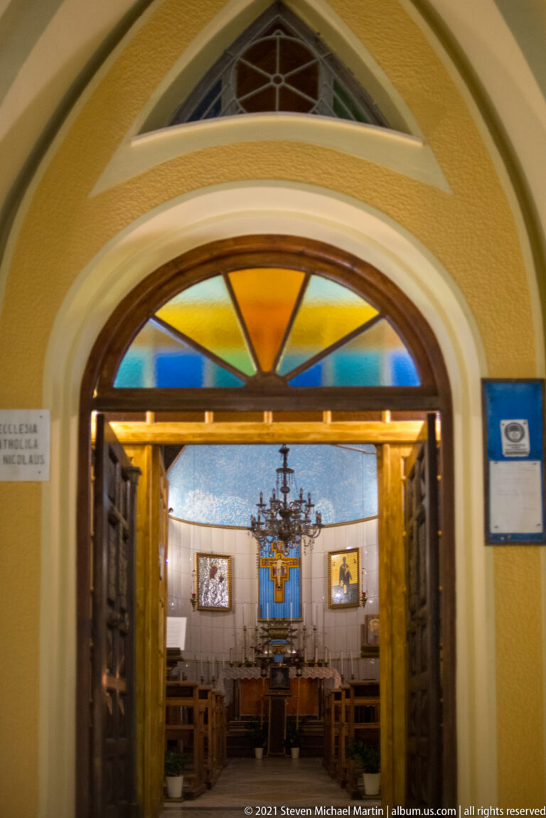 Open church doors beneath a fan-shaped colored-glass transom, framing the altar and cross inside.