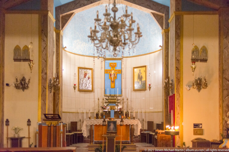 Christian sanctuary with altar, large crucifix on a blue panel, chandelier, framed icons, and marble-like walls.