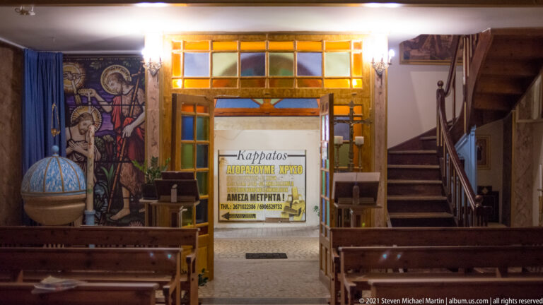 Interior with wooden benches, religious mural, colored glass, staircase, and a prominent sign reading Karpathos beyond the doorway.