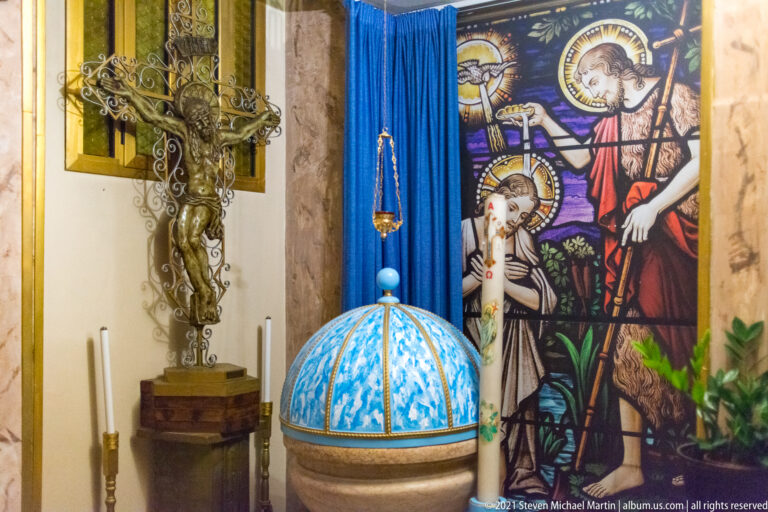 Blue-and-white covered baptismal font beside a colorful religious glass panel, metal cross, and hanging lamp.