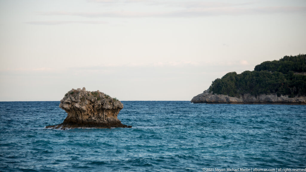 Single rugged rock formation rising from blue sea near a wooded coastal headland.