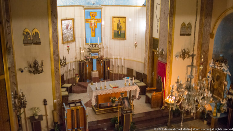 Elevated side view over a church sanctuary with altar, crucifix, apse seating, icons, and chandeliers.