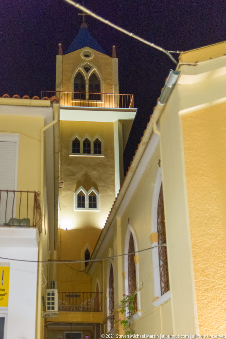 Illuminated church tower with pointed windows rising behind adjoining ochre rooflines against the night sky.
