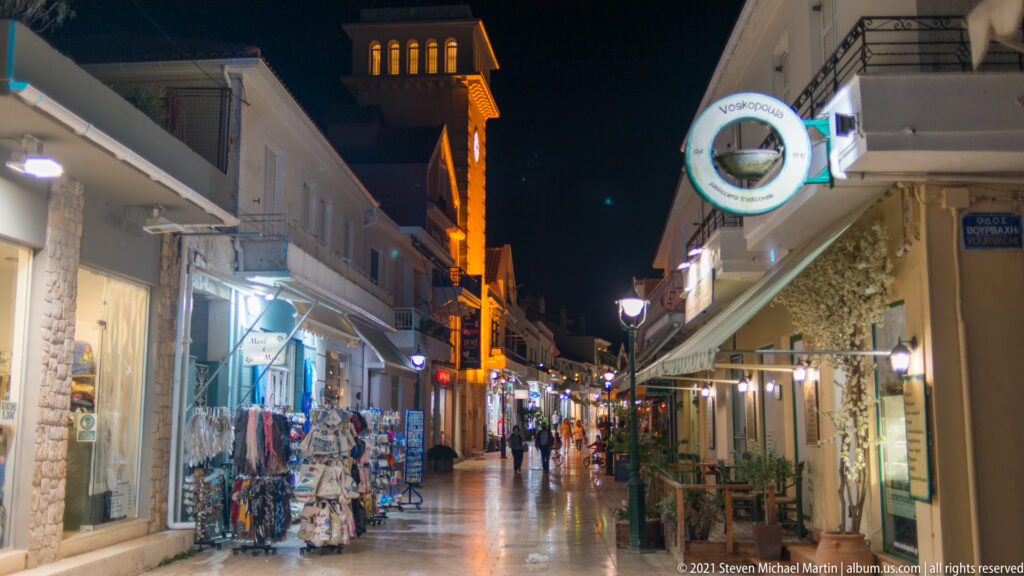 Nighttime pedestrian street lined with souvenir displays, shops, restaurants, walkers, and an illuminated tower in the distance.