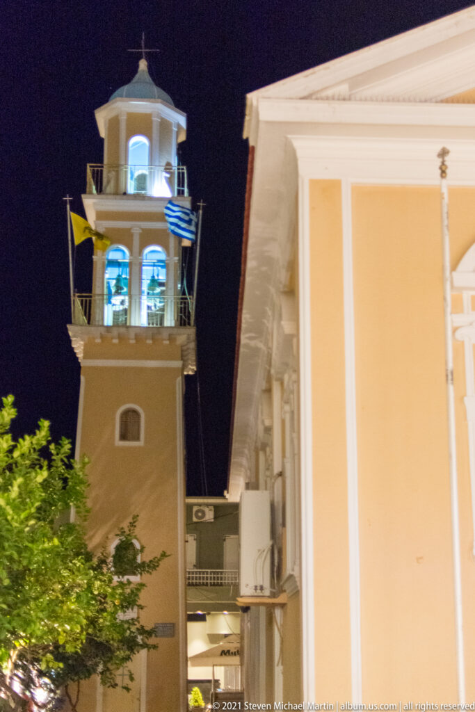 Square bell tower illuminated at night, with bright open belfry above trees beside a pale building.