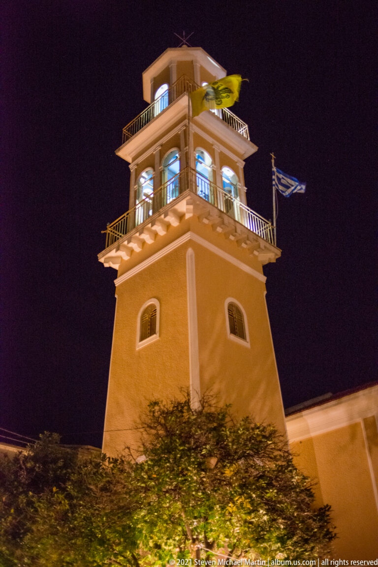 Low-angle night view of an illuminated square tower with open upper belfry, balconies, and a Greek flag.
