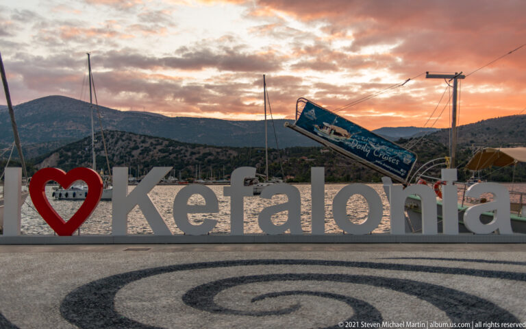 Large red heart and white Kefalonia letters on a waterfront promenade at sunset, with mountains and sailboat masts behind.