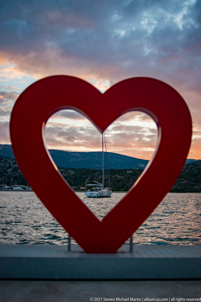 Large red heart sculpture framing a harbor, sailboat mast, distant shore, and colorful sunset sky.