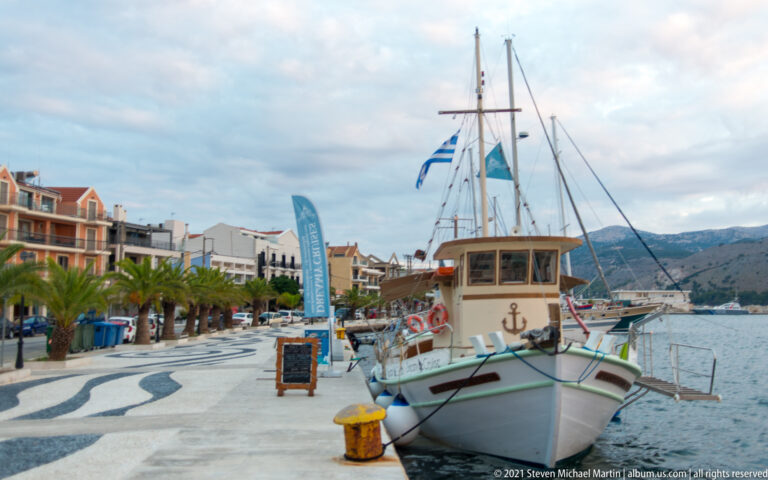 Small wooden passenger boat moored beside a patterned waterfront promenade with Greek flag, palms, buildings, and mountains.