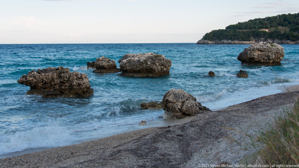 Pebbly shoreline with small breaking waves and several jagged rocks in shallow blue water, wooded headland beyond.