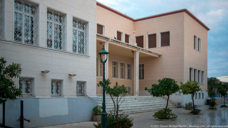 Large pale institutional building with broad entrance steps, rectangular windows, metal grilles, trees, and a traditional streetlamp.