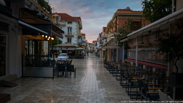 Pedestrian street lined with café tables, awnings, shops, and multistory buildings under an evening sky.