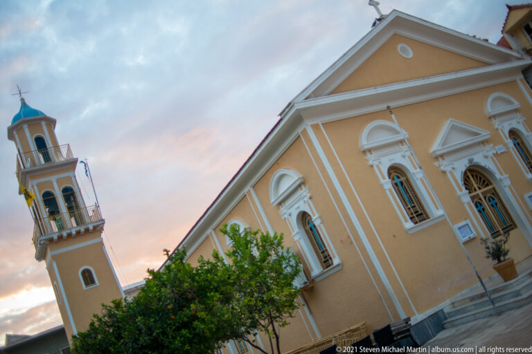Low-angle view of an ochre church and bell tower with white trim, arched windows, and a tree in front.