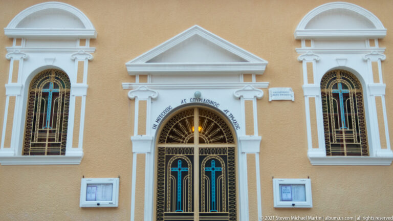 Centered church doorway with Greek lettering, flanked by two tall arched windows in an ochre-and-white façade.