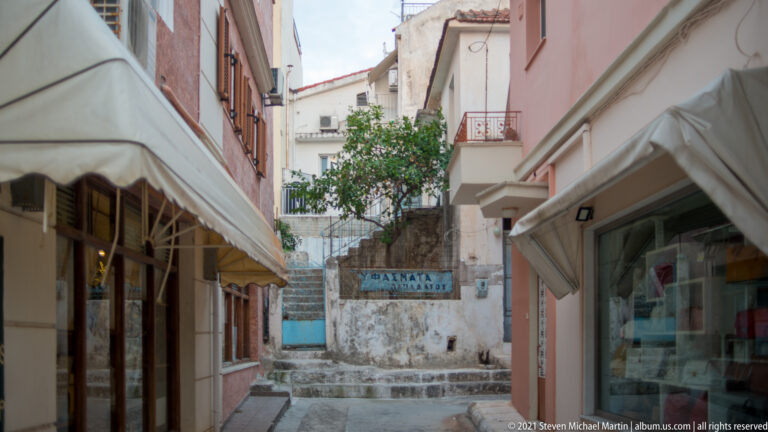 Narrow alley between pastel shopfronts leading to stone steps and a painted Greek sign on the end wall.