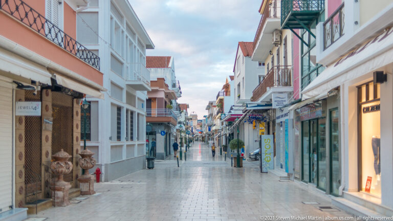 Pedestrian shopping street with pastel buildings, balconies, storefronts, signs, and a few people in the distance.