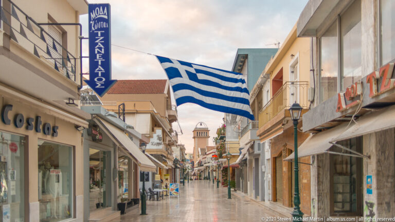 Large Greek flag hanging across a pedestrian shopping street with shops, awnings, tables, and a distant tower.