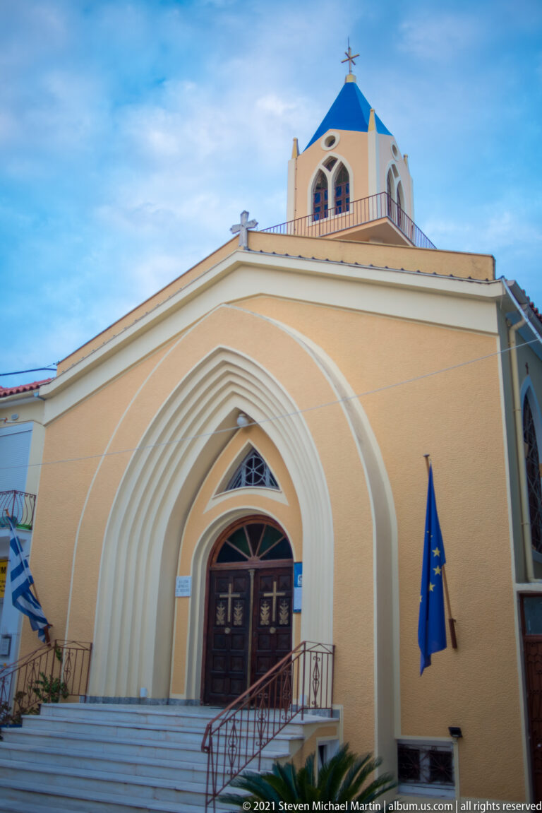 Front view of an ochre church with layered pointed-arch entrance, white steps, flags, and a small blue-roofed belfry.