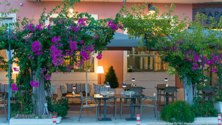 Outdoor café tables beneath dense purple bougainvillea beside a warmly lit building façade.