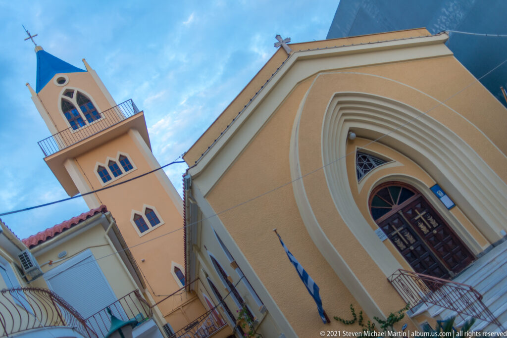 Low-angle oblique view of an ochre church with pointed-arch portal, square tower, balconies, and roof cross.