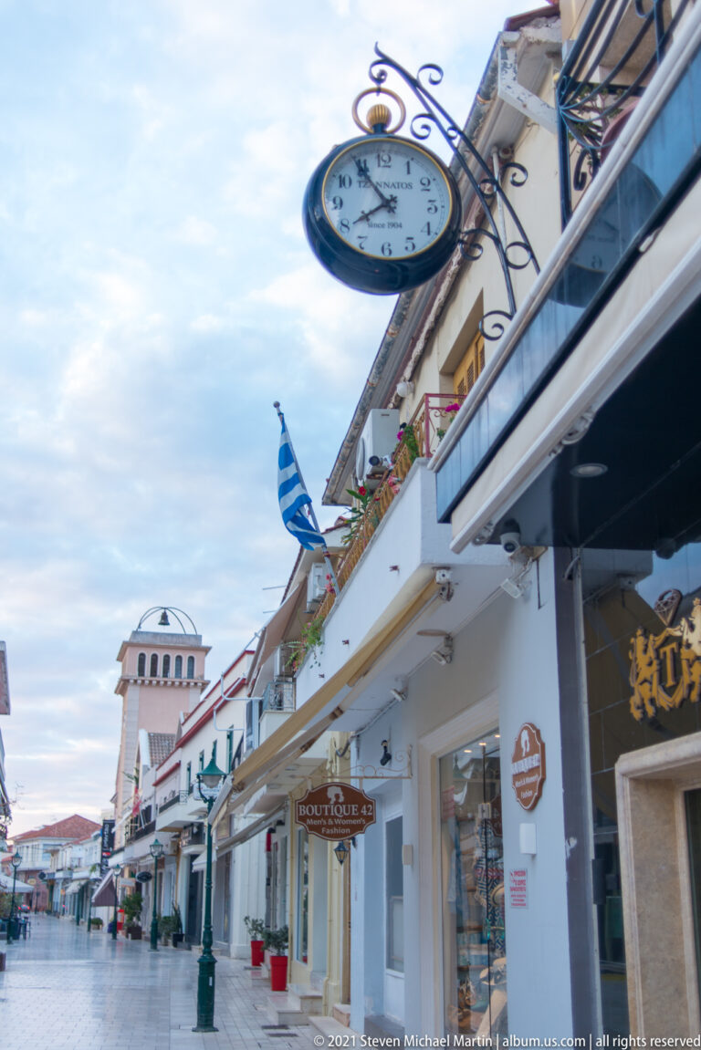 Round hanging street clock on an ornate bracket above shops, a Greek flag, and a distant tower.