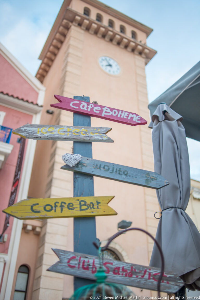 Colorful directional arrows for cafés and bars mounted below a pale clock tower and beside a folded umbrella.