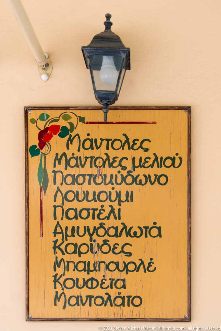 Wooden sign with multiple lines of green Greek lettering and a painted red-and-green parrot beneath a black lantern.