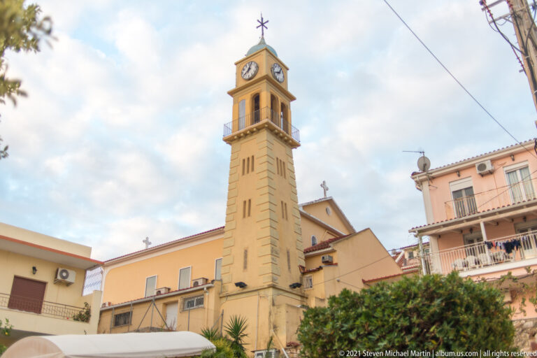 Slender ochre church clock tower with belfry, clock face, and cross rising above nearby houses and trees.