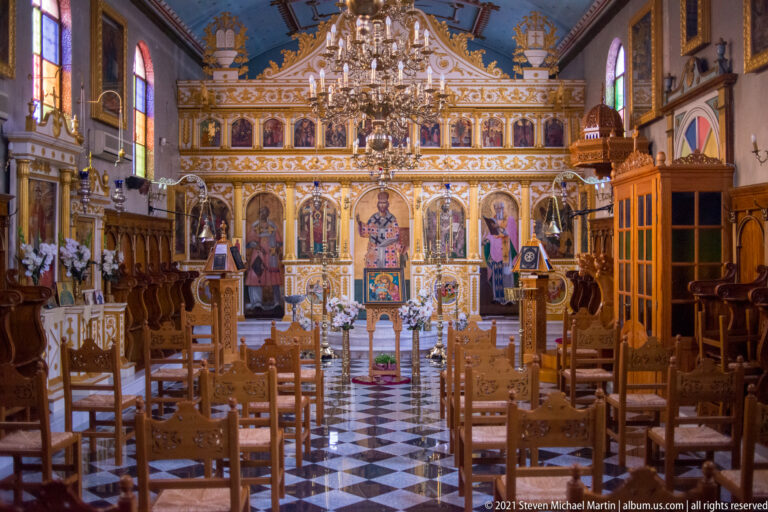 Orthodox church nave with black-and-white floor, wooden chairs, chandeliers, and an ornate gilded iconostasis.