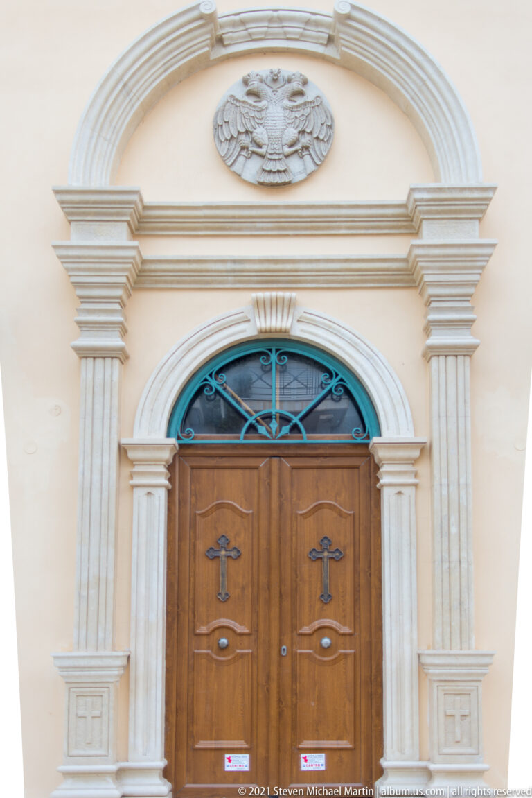 Double wooden church doors with cross motifs set in a pale arched portal with pilasters, transom grille, and carved crest.