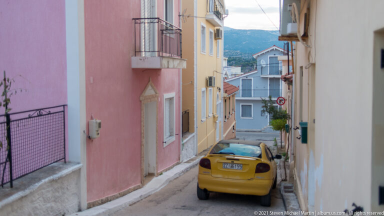 Narrow residential lane between pink and yellow houses with a parked yellow car and green hillside visible beyond.