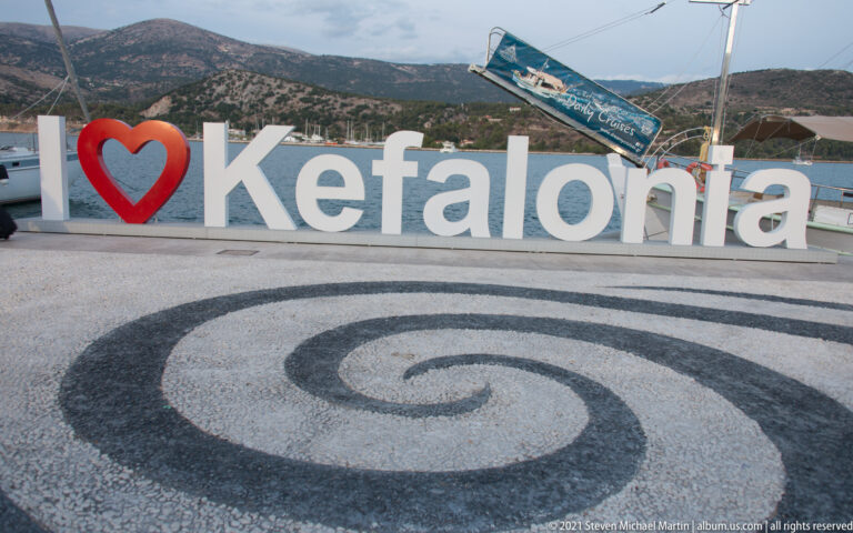 Large white Kefalonia letters with red heart behind a black-and-white spiral pavement design at the waterfront.
