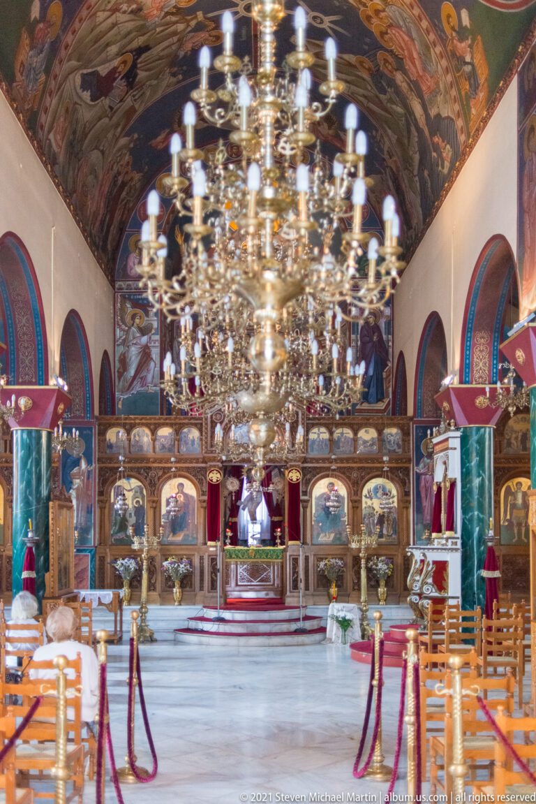 Large Orthodox church nave with several brass chandeliers, rows of chairs, painted vaults, and iconostasis beyond.