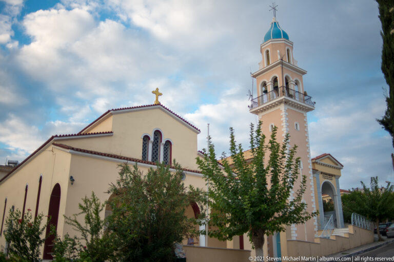 Side view of a pale Orthodox church with red roof trim, trees, and a tall bell tower capped in blue.