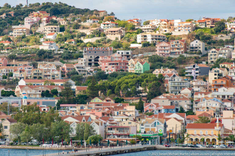 Pastel houses and apartment buildings descend a green hillside toward a waterfront road and blue water.