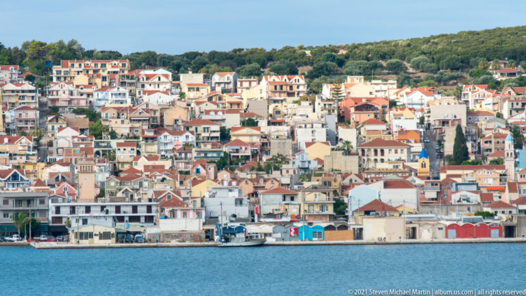 Compact coastal town of pale and pastel buildings with red roofs lining the shore and climbing a wooded hillside.