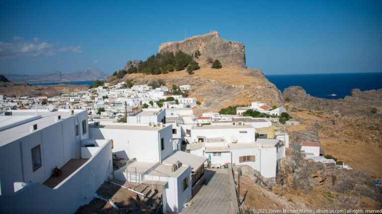 Whitewashed Lindos houses below a steep rocky acropolis crowned by defensive walls, with the Aegean Sea beyond.