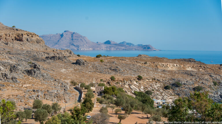 Dry limestone valley near Lindos with olive trees, scattered buildings, sea, and hazy mountains in the distance.