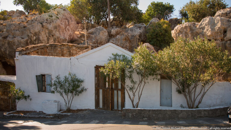 Small whitewashed Lindos house with timber doors and trees, built directly against a rough rocky hillside.