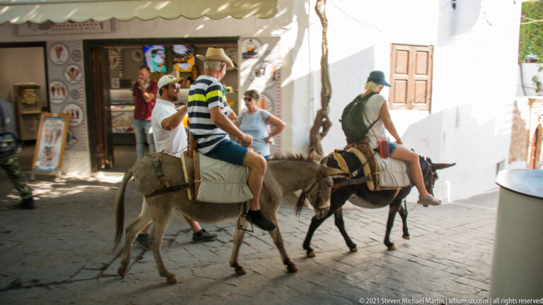 Two visitors ride saddled donkeys along a narrow whitewashed shopping lane in Lindos as pedestrians pass nearby.