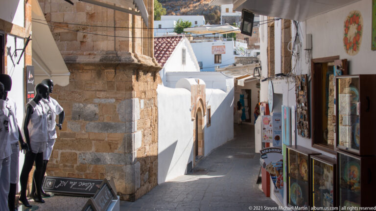 Narrow Lindos shopping lane between a stone building and whitewashed storefronts, with pedestrians and merchandise displays.