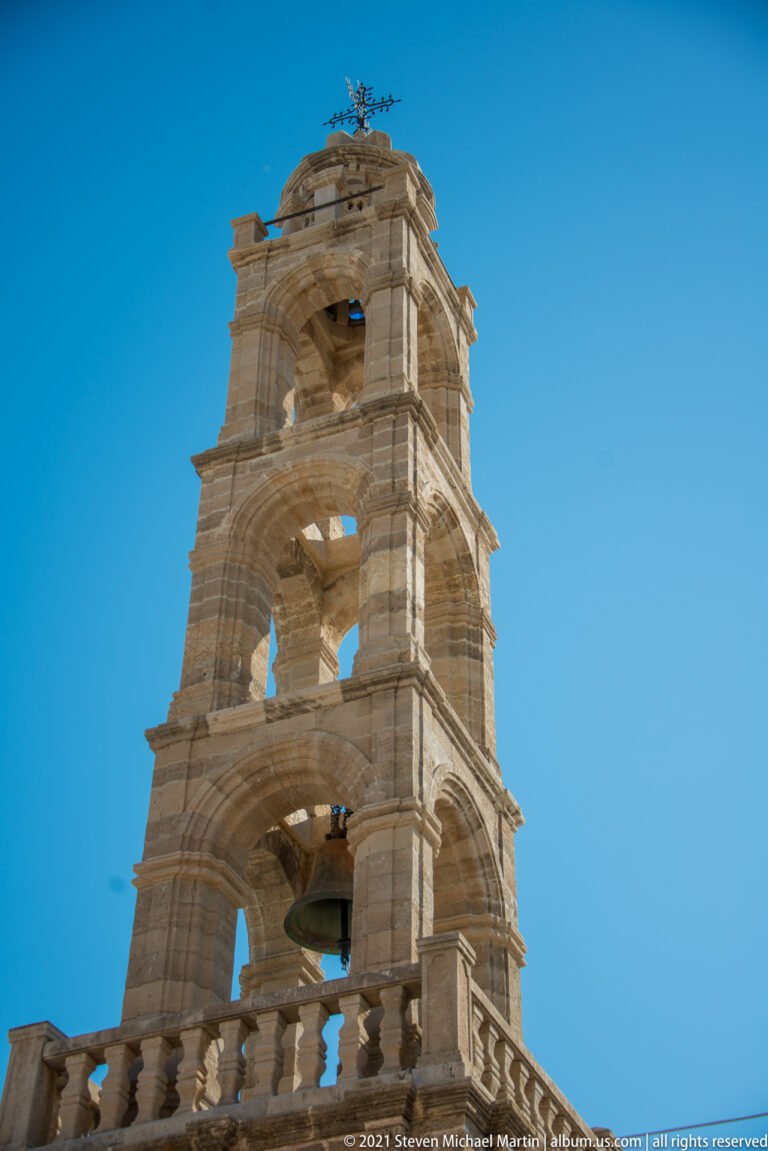 Tall stone church bell tower in Lindos with stacked arched openings and bells, seen from below against a blue sky.