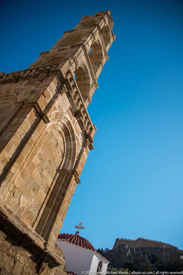 Oblique upward view of a stone church façade and bell tower in Lindos, with arched details against a clear blue sky.