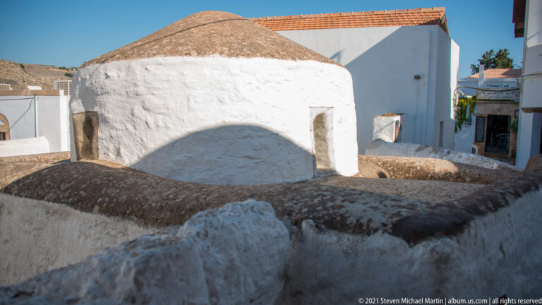 Small whitewashed domed structure with a rectangular opening, surrounded by closely packed roofs and walls in Lindos.