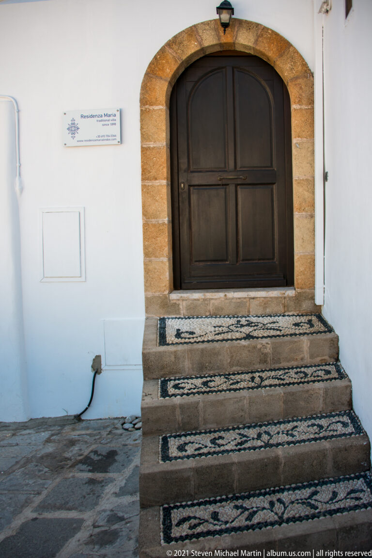 Dark wooden double door in a stone arch above black-and-white pebble-mosaic steps and whitewashed walls in Lindos.